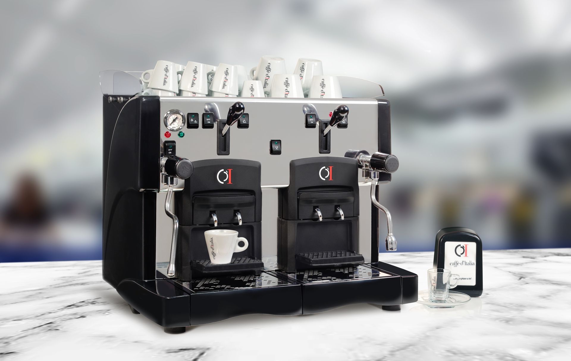 Macchina da caffè doppia nera con tazze sulla parte superiore e un piccolo display per smartphone accanto, su un piano in marmo.