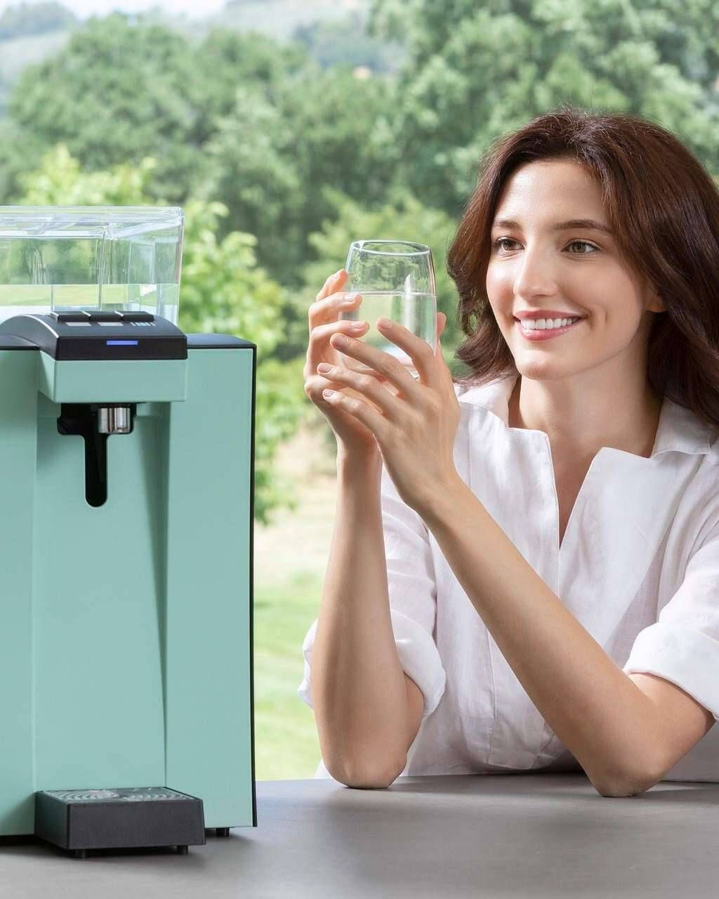 Una donna sorride mentre tiene in mano un bicchiere accanto a un distributore d'acqua verde menta all'aperto.