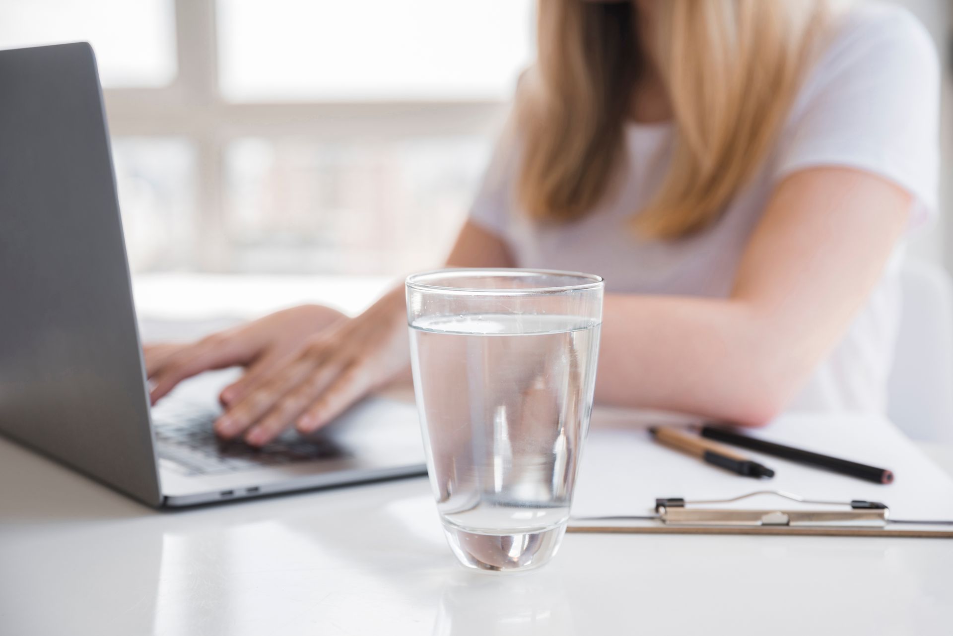 Una persona digita su un computer portatile accanto a un bicchiere d'acqua e una penna su una scrivania luminosa.
