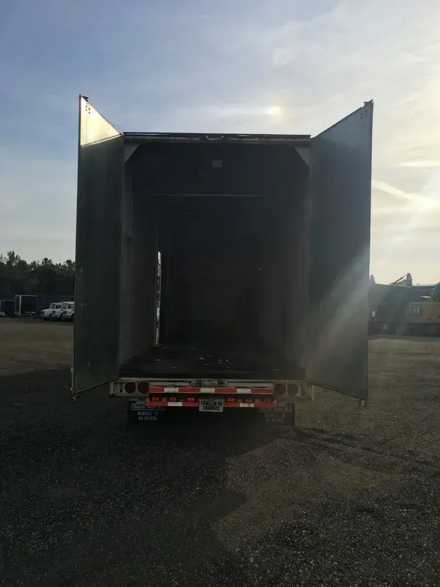 Open cargo trailer, doors ajar, parked on gravel. Back, red and white lights. Outdoors, sunny.