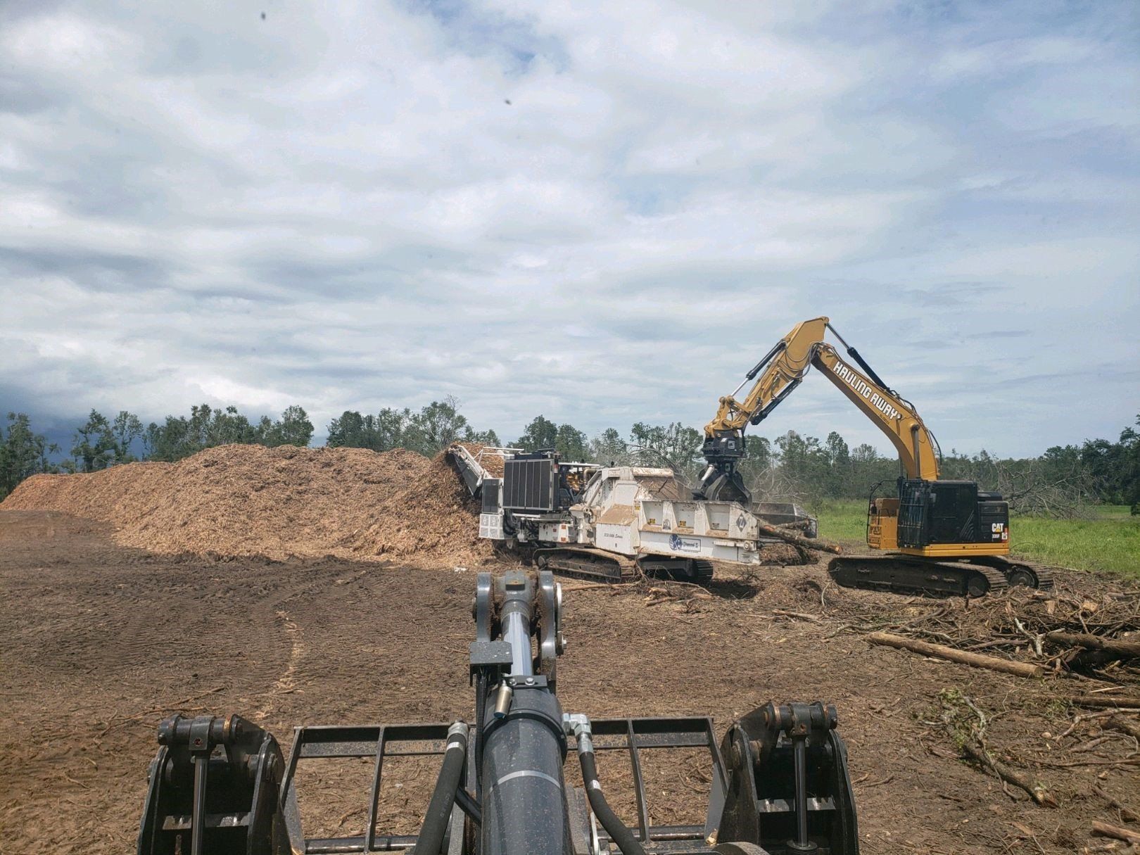 A large excavator is moving a pile of wood in a field.