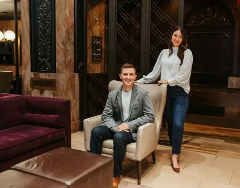 A man is sitting in a chair next to a woman standing next to him.