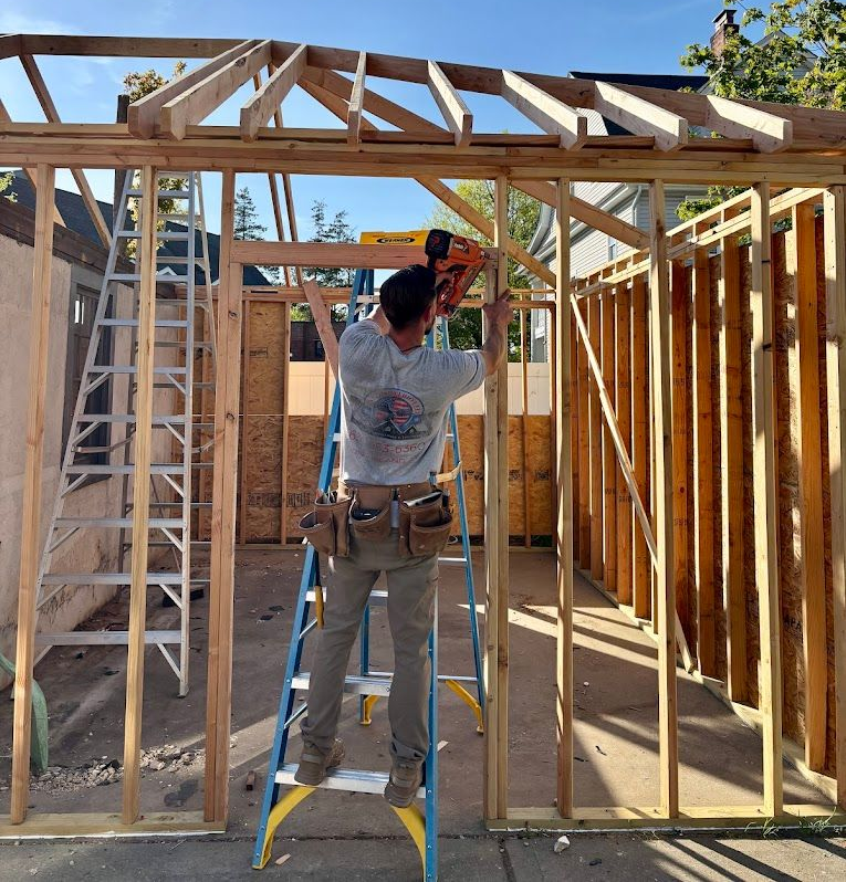 Construction worker framing a building with a nail gun, standing on a blue ladder. Sunlight.