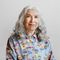 Woman with long gray hair smiles, wearing a blue floral shirt, against a light gray backdrop.