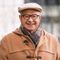 Smiling man wearing glasses, beige cap, coat, and scarf; outdoors.