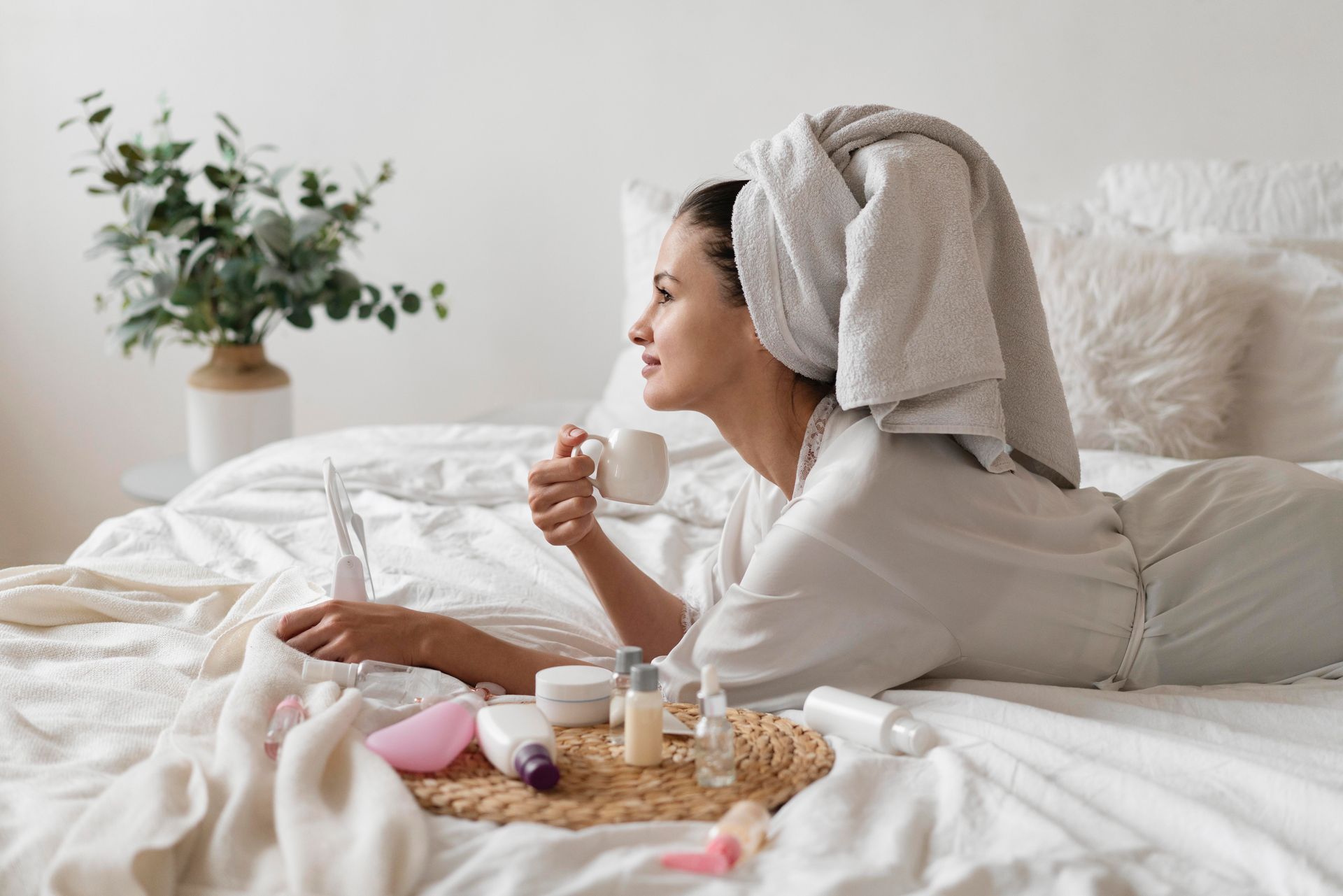 Mulher de roupão na cama realizando sua rotina de autocuidado noturna com produtos de beleza.