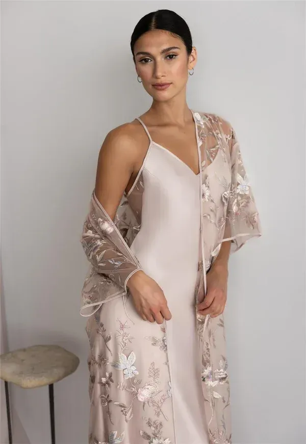 Woman wearing a satin slip dress and floral embroidered shawl, standing near a stool.