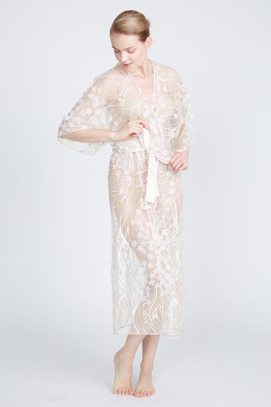 Woman in sheer, floral lace robe, tied at the waist, looking down. White background.