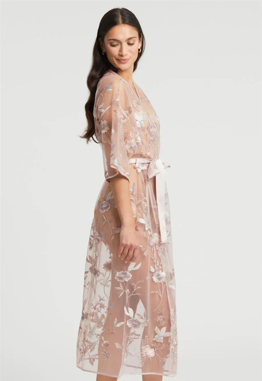 Woman in a sheer, floral-embroidered blush robe, tied with a ribbon, looking down.