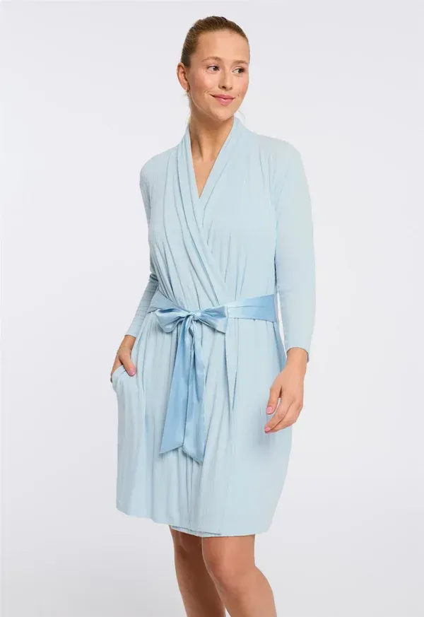 Woman in light blue robe with a tie, standing indoors with a slight smile.