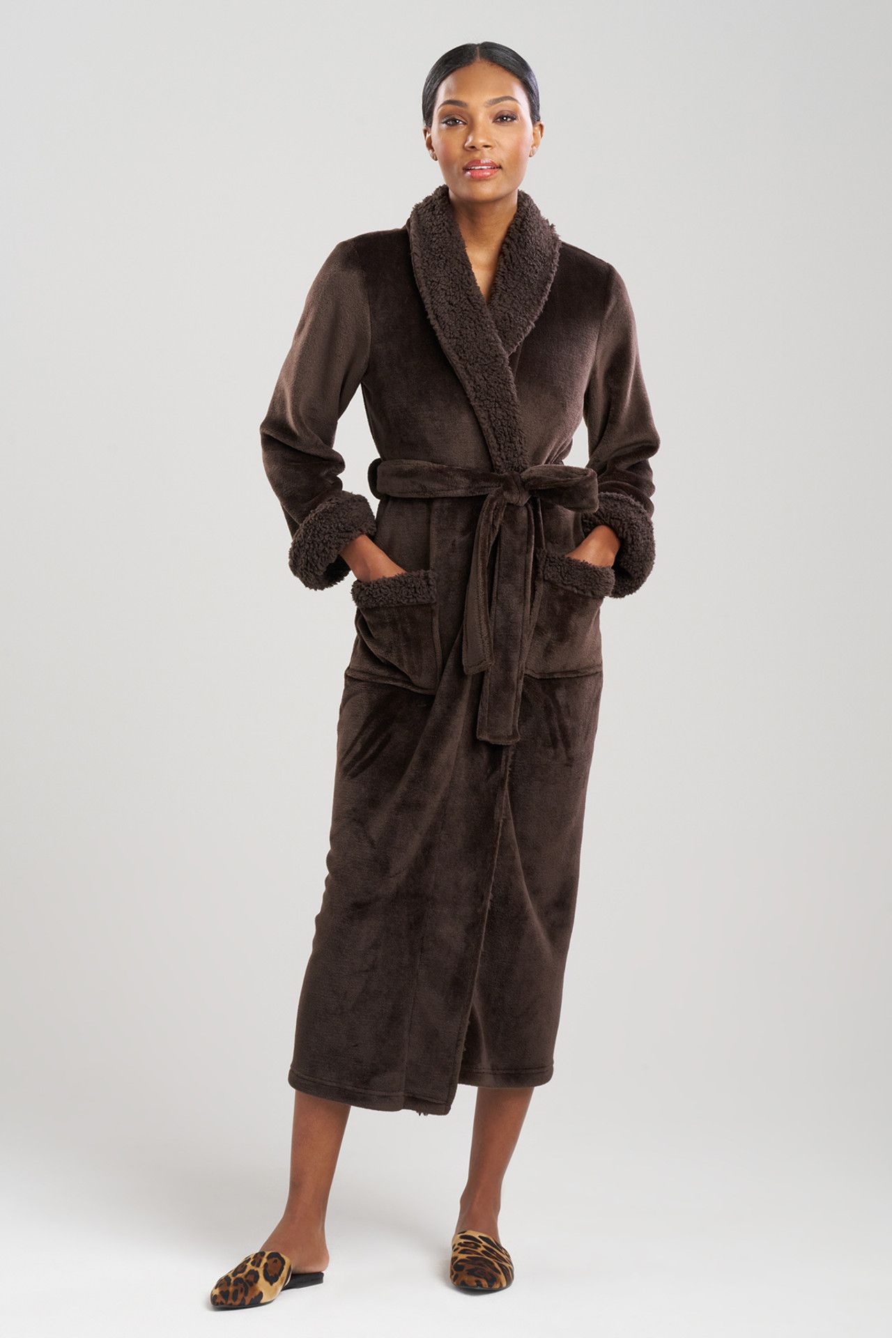 Woman in a brown plush robe, standing, hands in pockets. Neutral setting, wearing leopard print slippers.