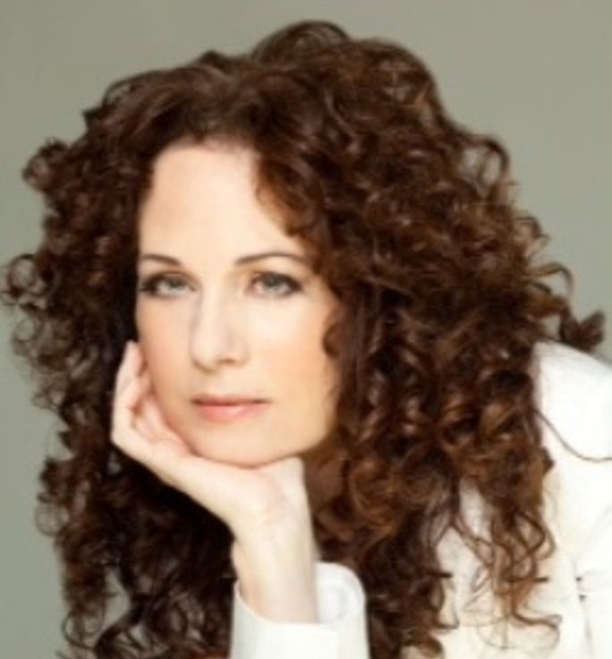 Woman with curly auburn hair, resting her head on her hand, gazing forward.