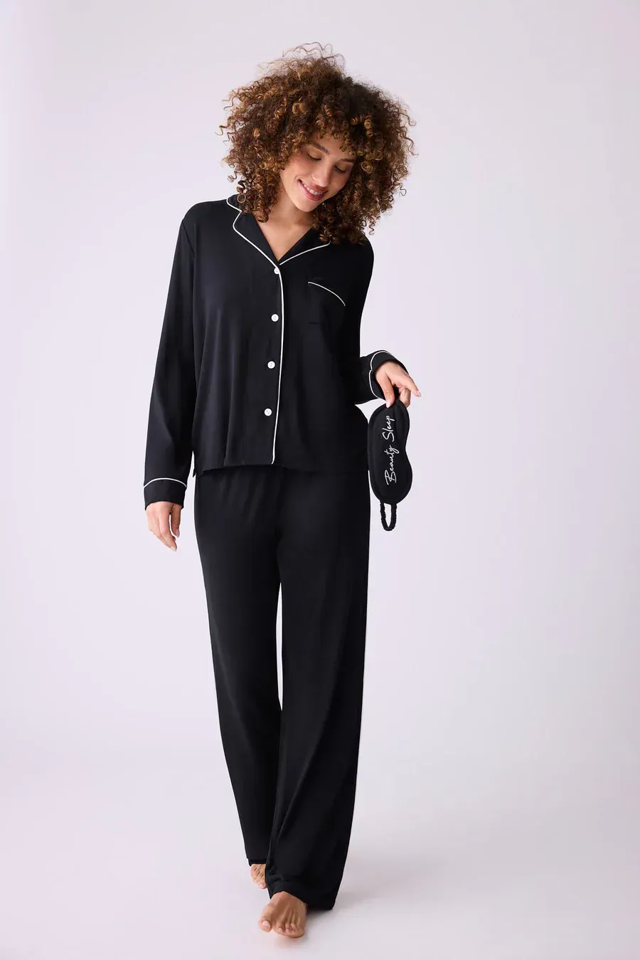 Woman in black pajamas holding a sleep mask, smiling, standing against a white backdrop.