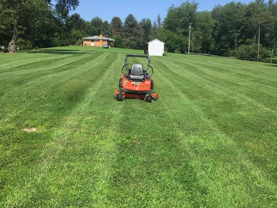 An orange riding lawn mower sitting in the center of a freshly mown, striped grass field with a house in the background.