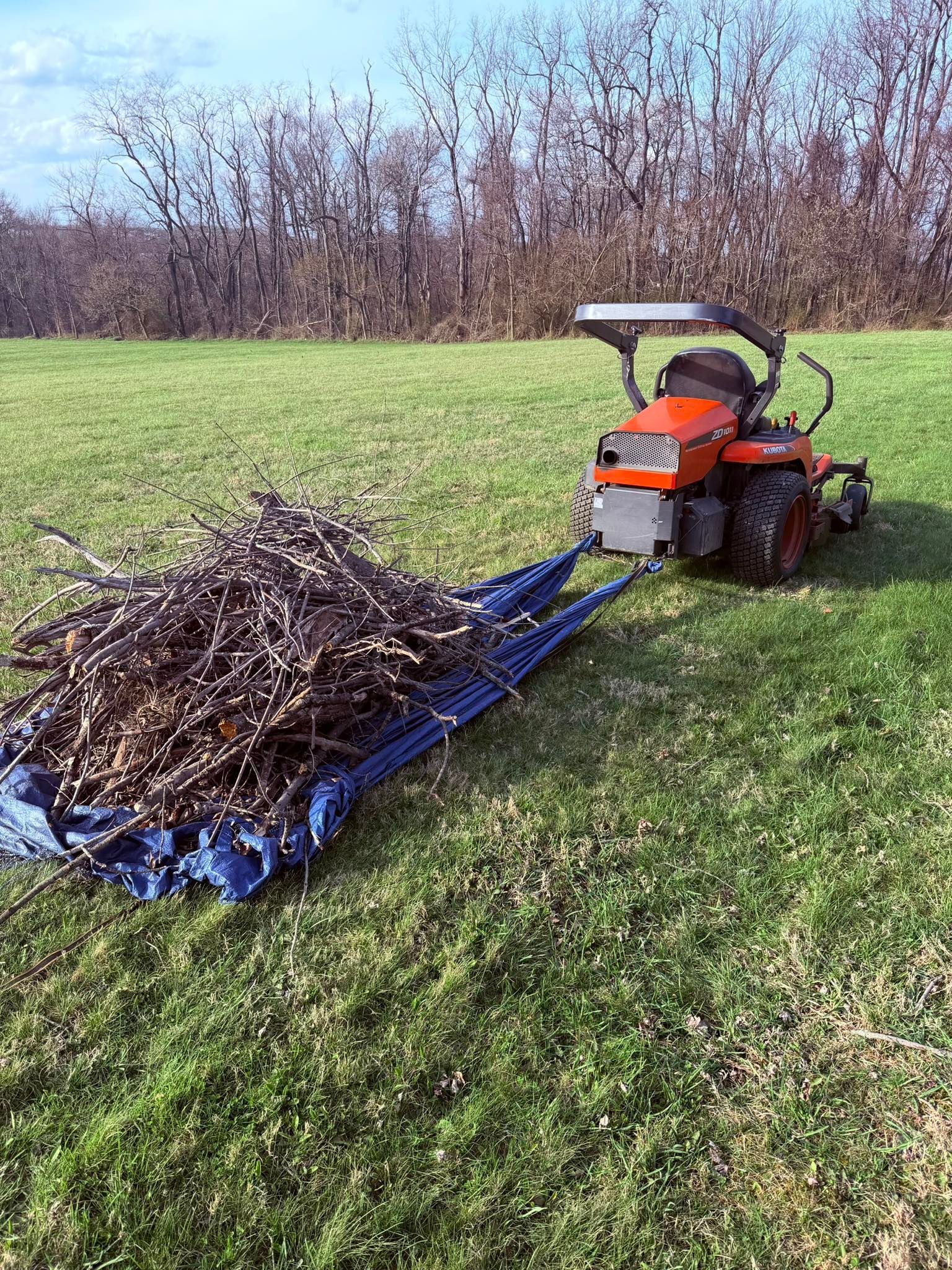 An orange tractor pulls a large blue tarp filled with branches across a grassy field under a bright, sunny sky.