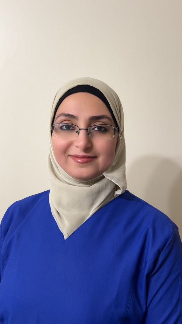 A woman wearing a hijab and glasses is standing in front of a white wall.