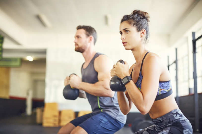 A man and a woman are squatting with kettlebells in a gym.