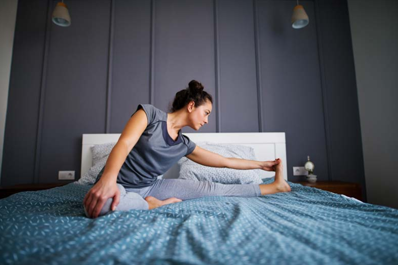 A woman is sitting on a bed stretching her legs.