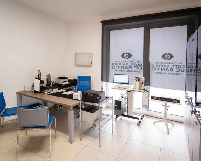 Uno studio medico con una scrivania, due sedie blu e attrezzature oftalmiche davanti a una finestra con tende di marca.