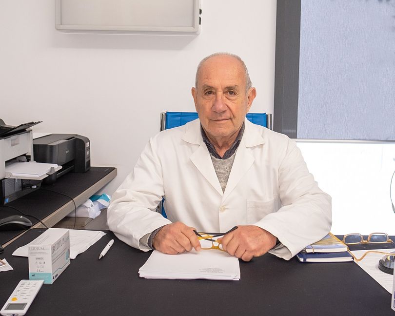 Un professionista sanitario in camice bianco siede a una scrivania con delle scartoffie e una penna in un ambulatorio.