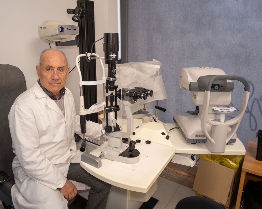 Un oftalmologo siede in una sala visite accanto a una lampada a fessura e a un apparecchio diagnostico automatizzato posizionato su un tavolo regolabile.