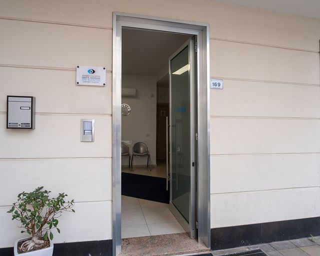 Una porta d'ufficio aperta con telaio in metallo, un'insegna, una cassetta delle lettere e una piccola pianta in vaso sulla parete chiara.