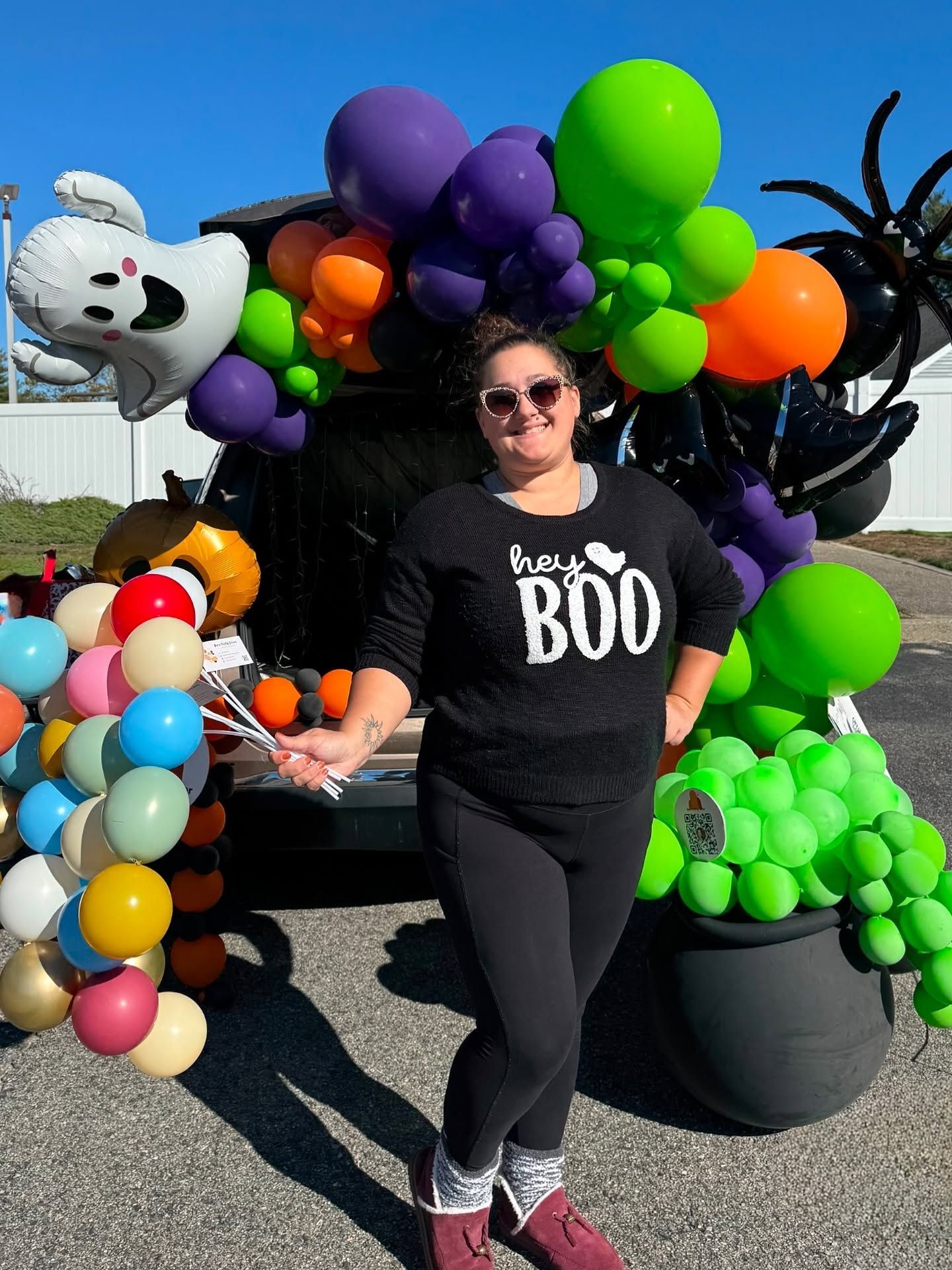 Person in black “BOO” shirt posing with Halloween balloons and decorations outdoors