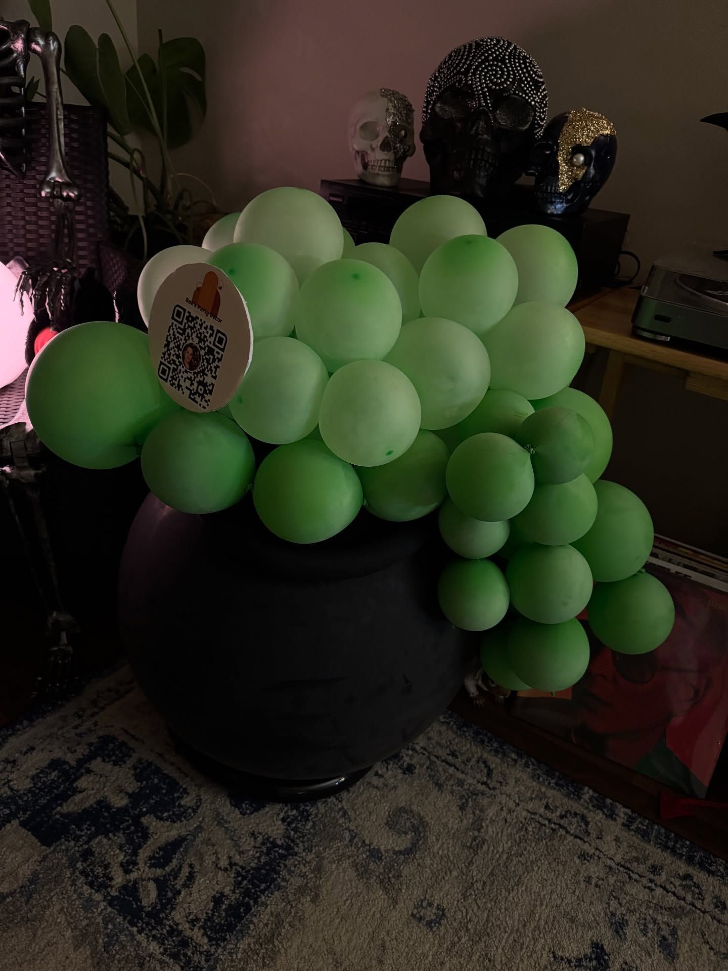 Green balloons clustered in a black bucket, with a decorated balloon near the front, in a dim room.