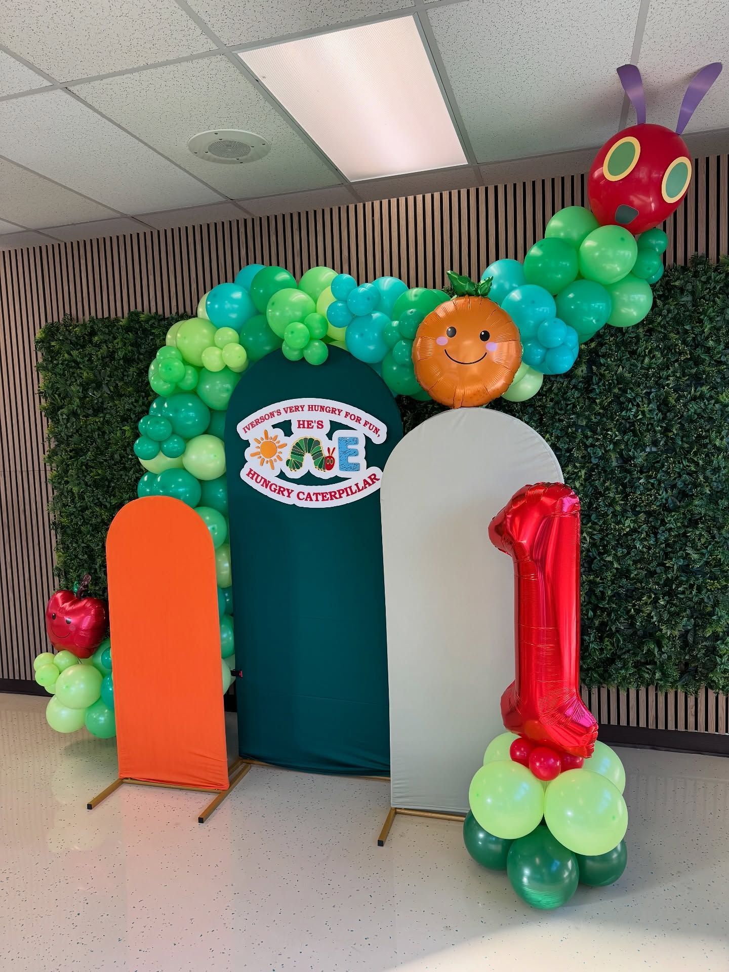 Colorful balloon arch frames a party entrance with a teal backdrop and orange “1” balloon.