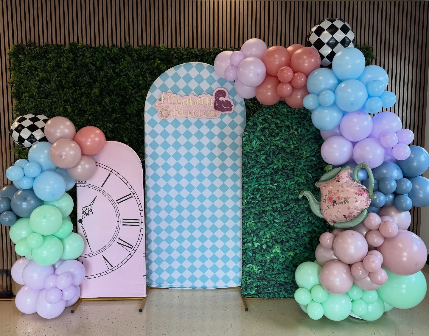 Pastel balloon arch around decorative panels, including a pink clock backdrop and blue checkered display.