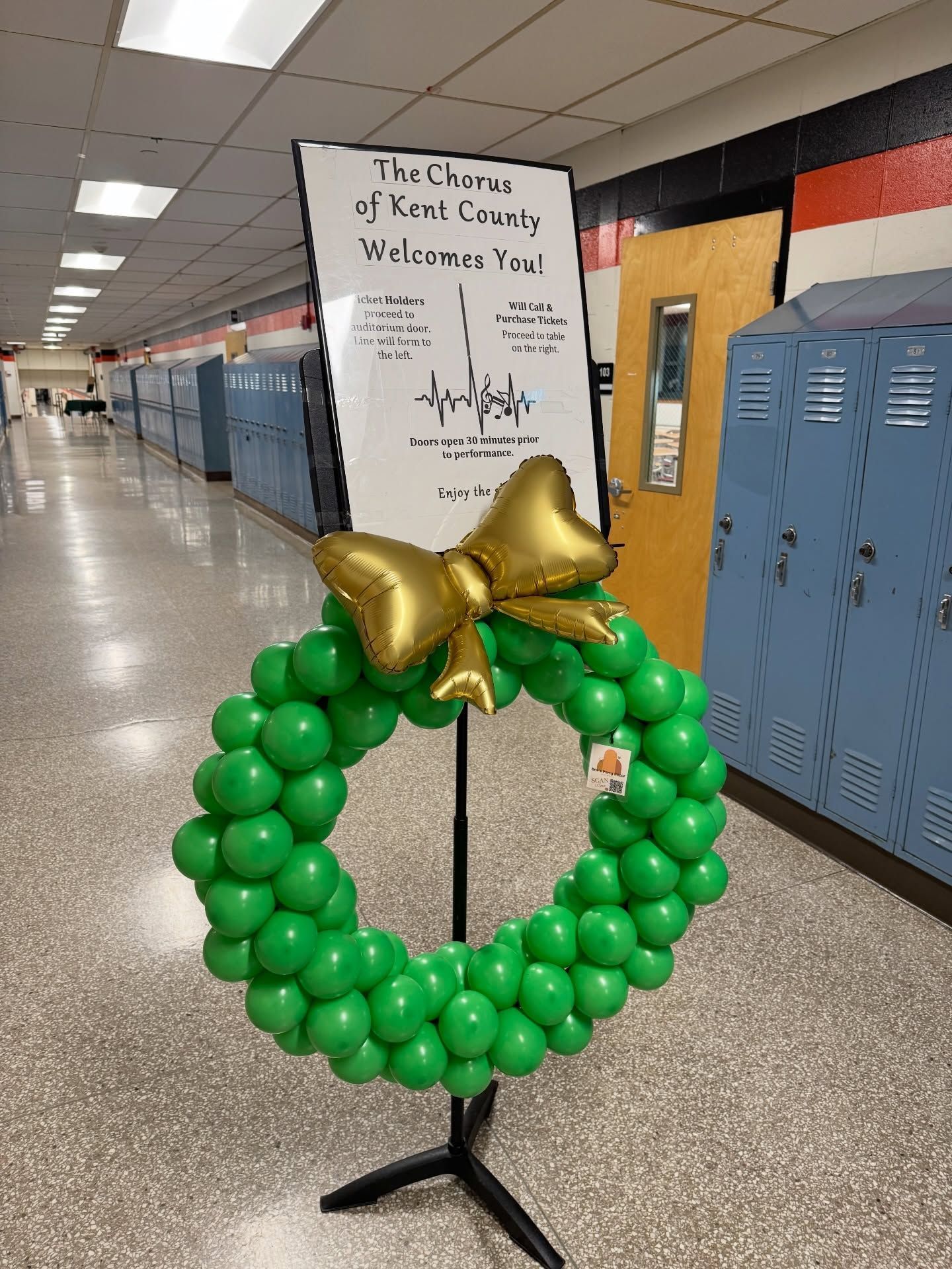 Green balloon wreath with gold bow on a stand in a school hallway beside lockers and a welcome sign