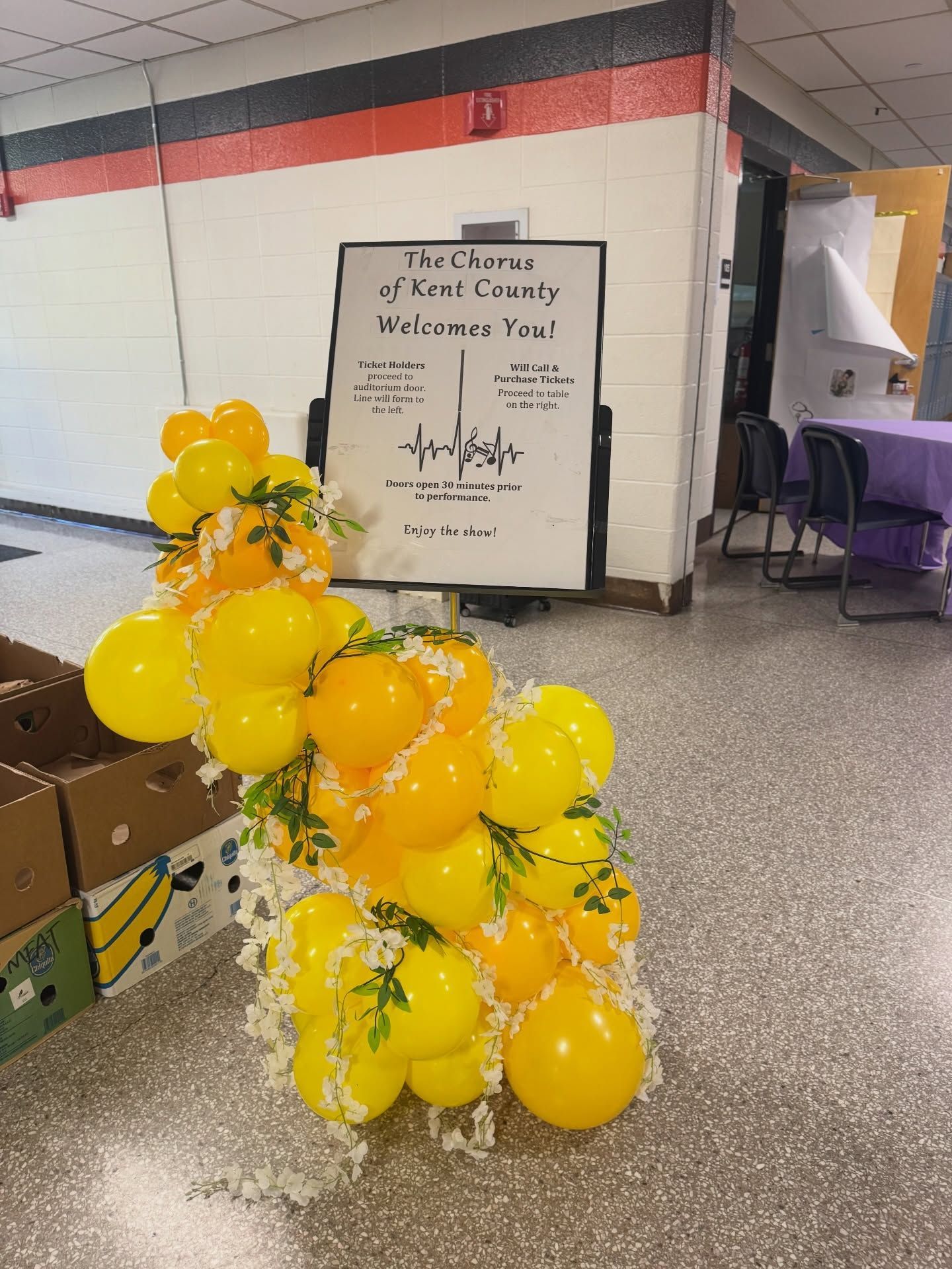Yellow balloon bouquet beside a sign in a school hallway with purple chairs and a black-and-red wall stripe