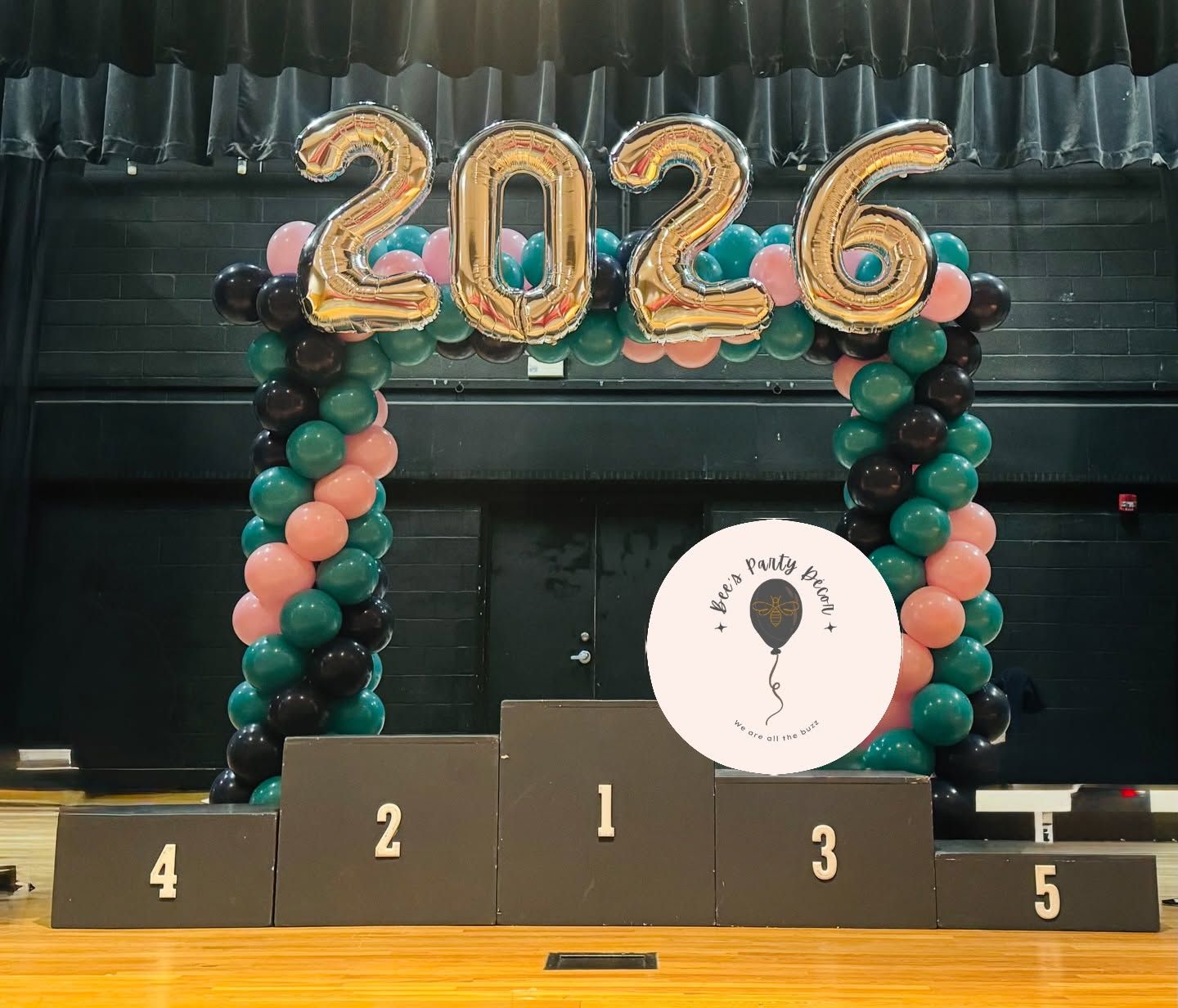 Gold “2026” balloons over a podium with black and teal balloon columns in a gymnasium.