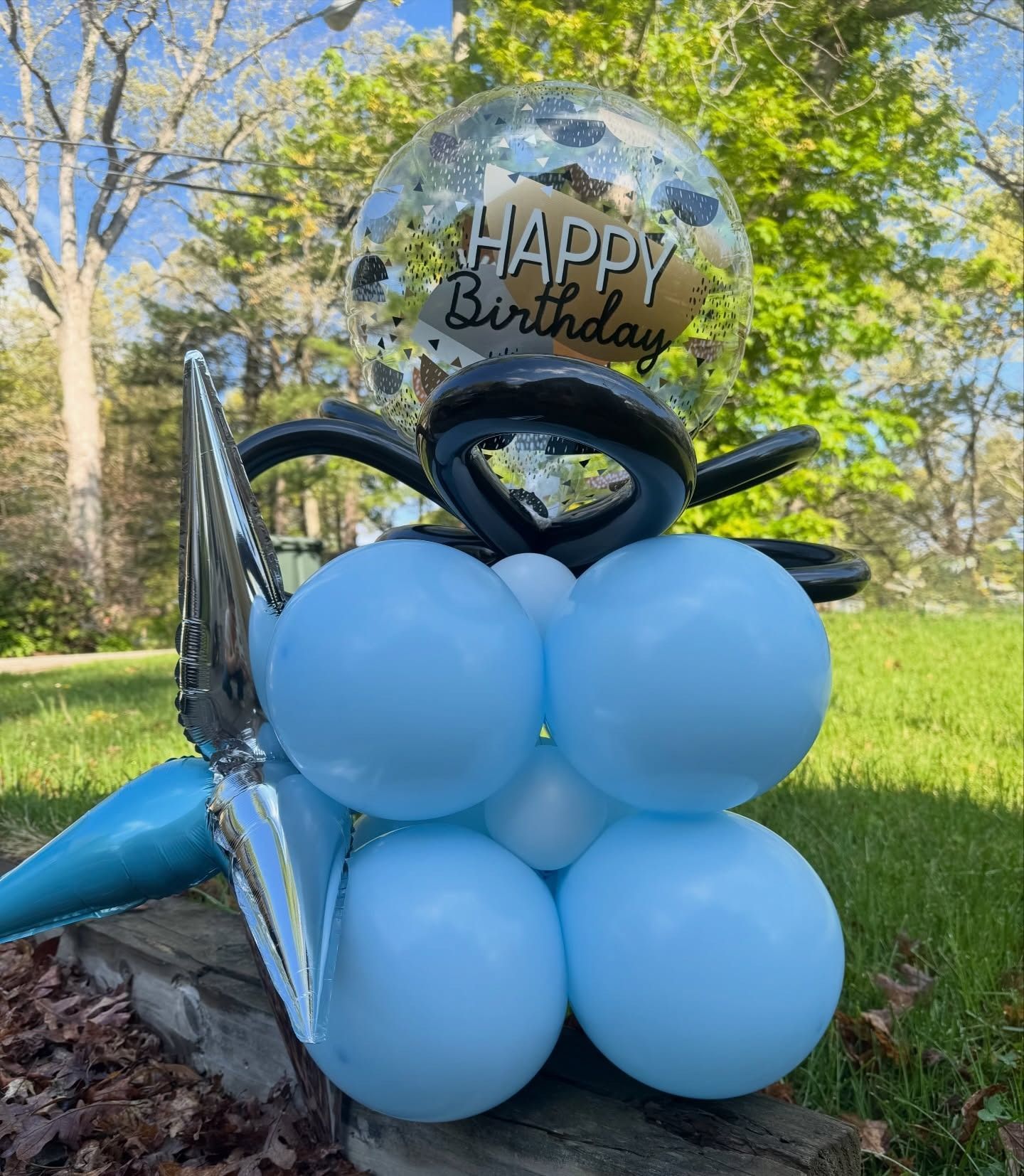 Birthday balloon sculpture with blue balloons and a clear “Happy Birthday” balloon outdoors on grass.