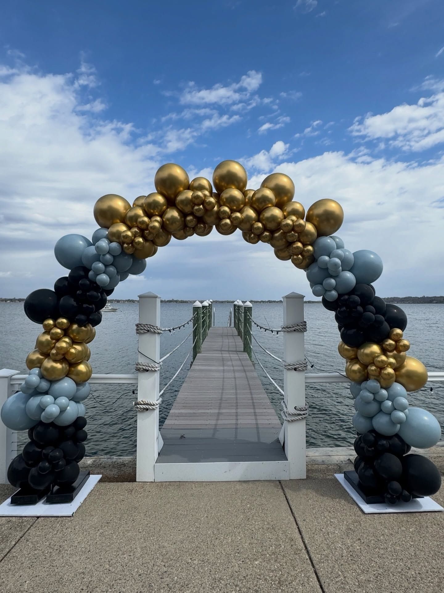 Black, gold, and silver balloon arch framing a white aisle entrance on a seaside walkway.