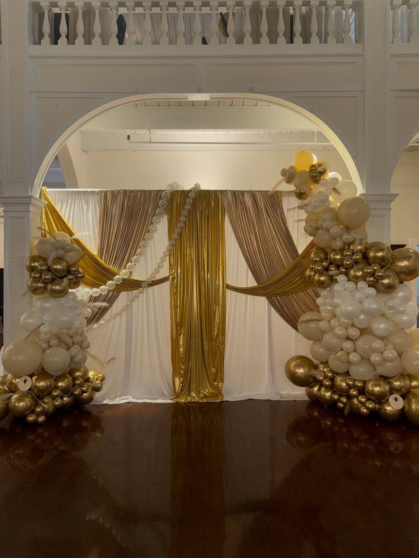 Gold and white balloon arch framing a draped event backdrop in a grand hall