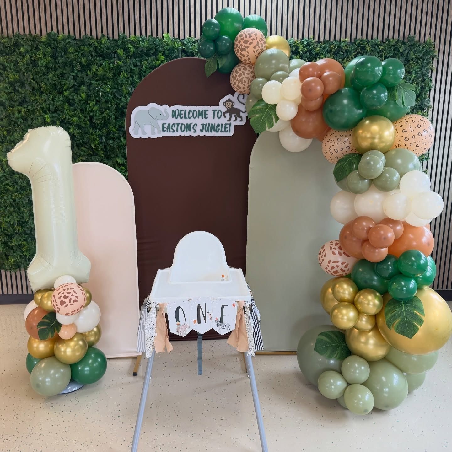 Gold, green, and white balloon display with a “1” and “ONE” chair in front of a welcome sign backdrop