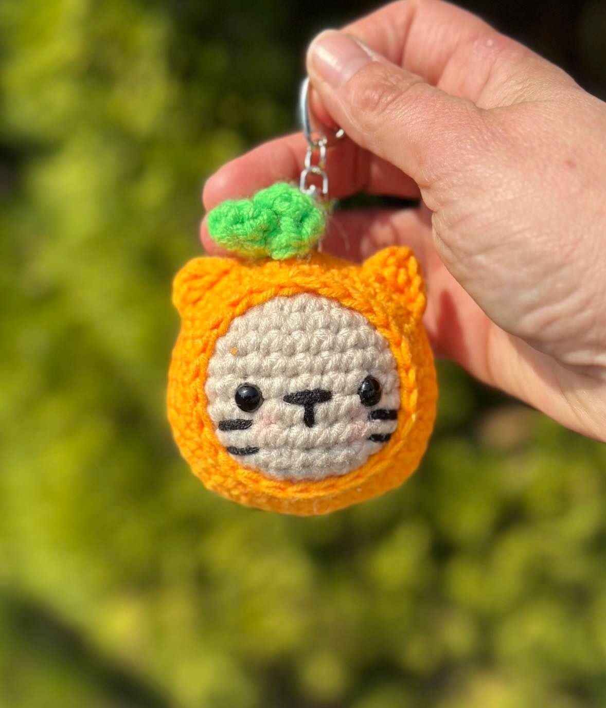 Crocheted orange cat keychain with green leaf held in a hand against a blurred green background.