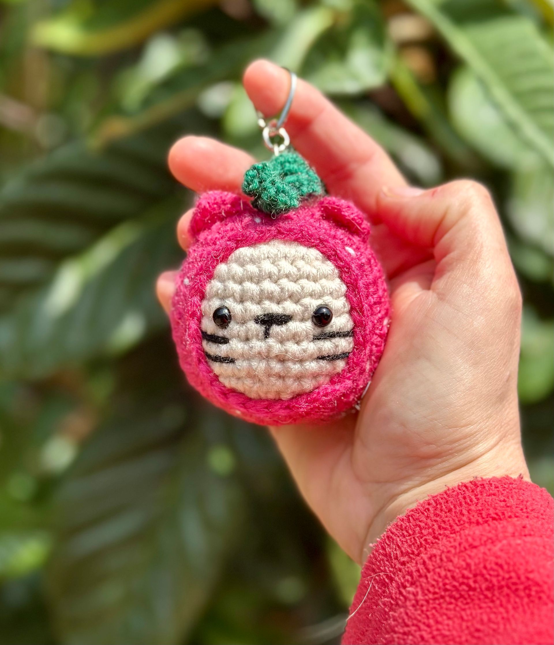 Crocheted pink cat keychain with green top, held in hand against a blurred green backdrop.
