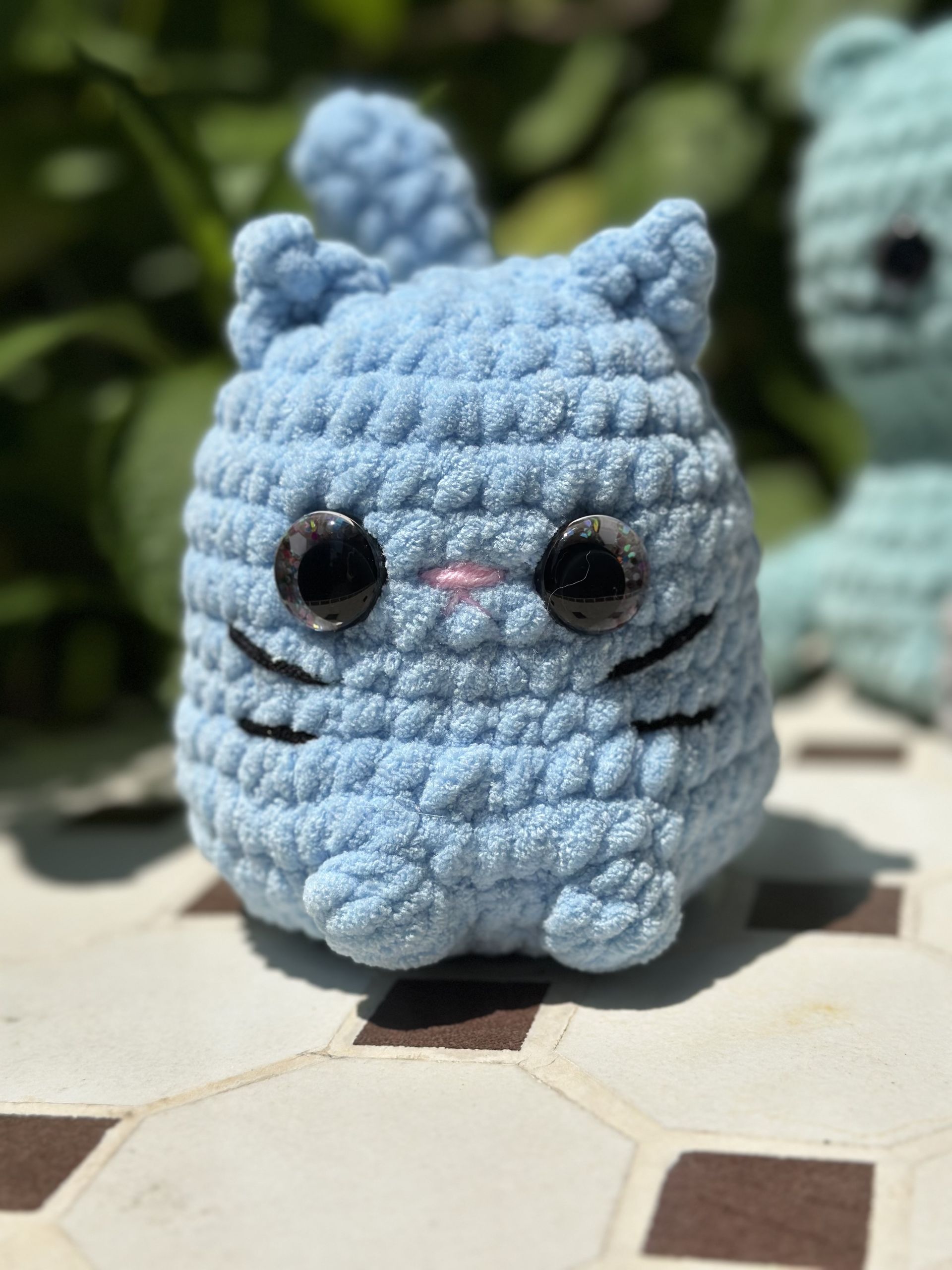 Crocheted blue cat plush held in a hand; features large black eyes, a pink nose, and a curled tail.