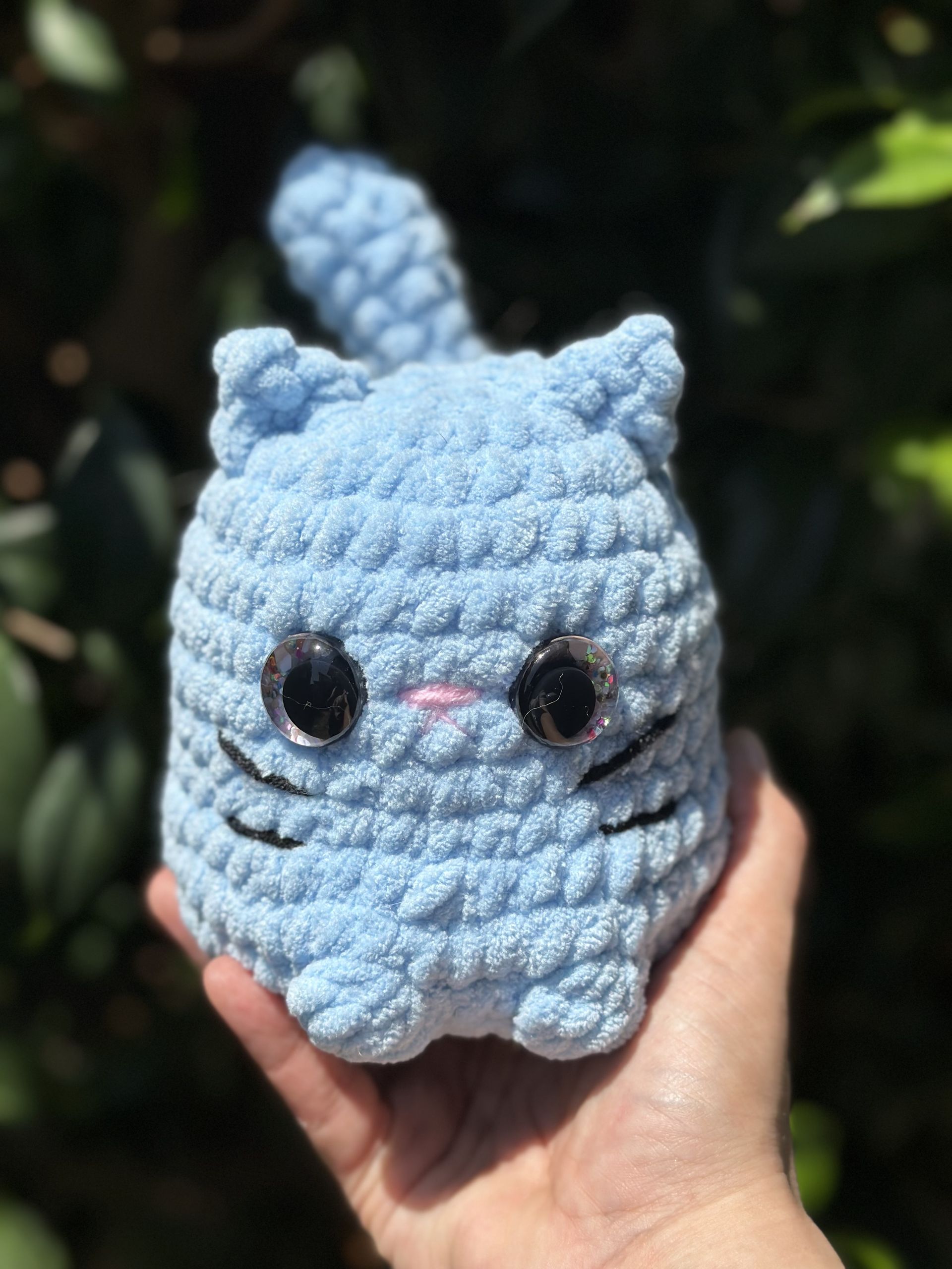 Crocheted blue cat plush held in a hand; features large black eyes, a pink nose, and a curled tail.