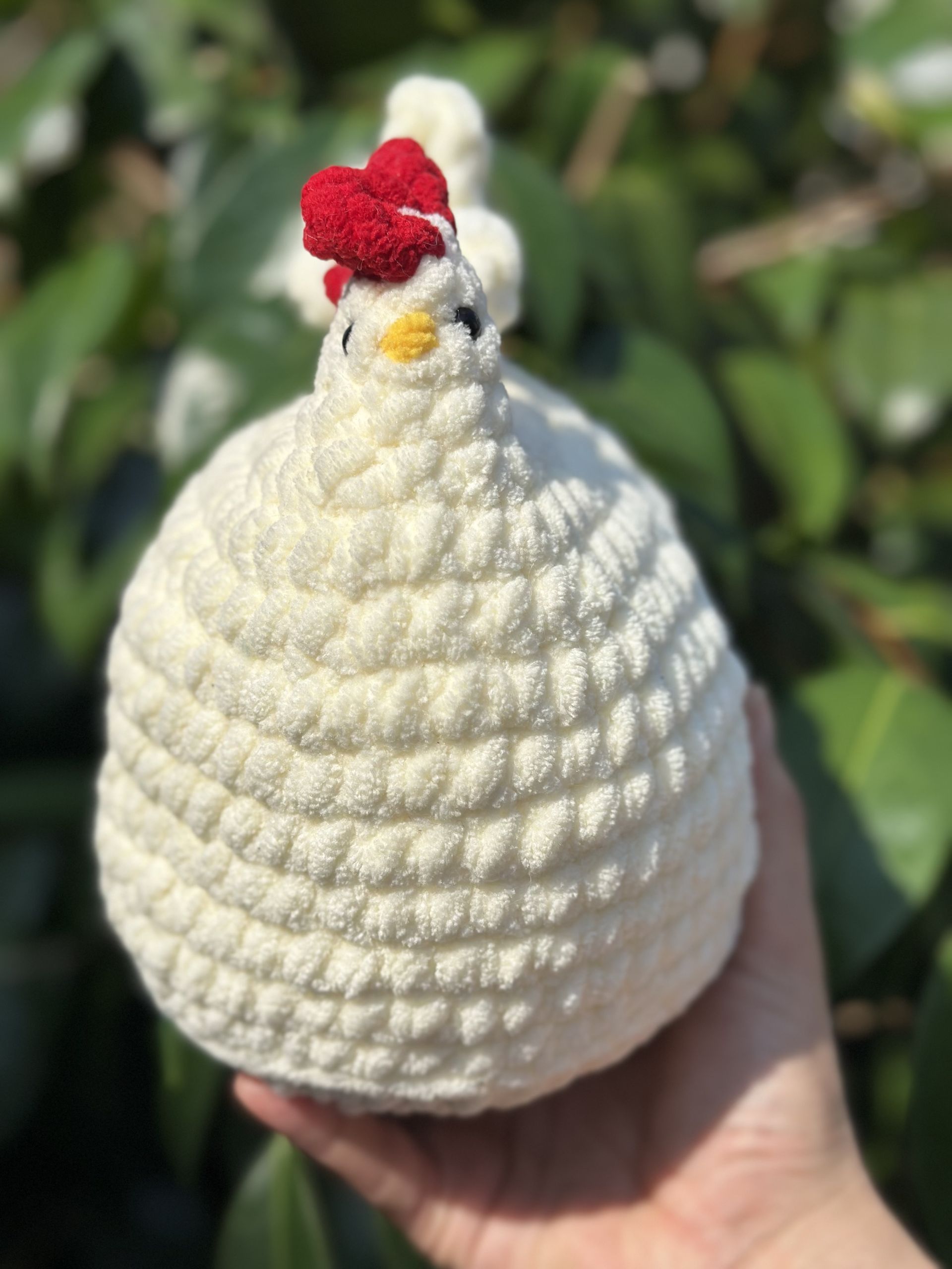 Crocheted white chicken figurine with red comb, held in hand outdoors, surrounded by green foliage.