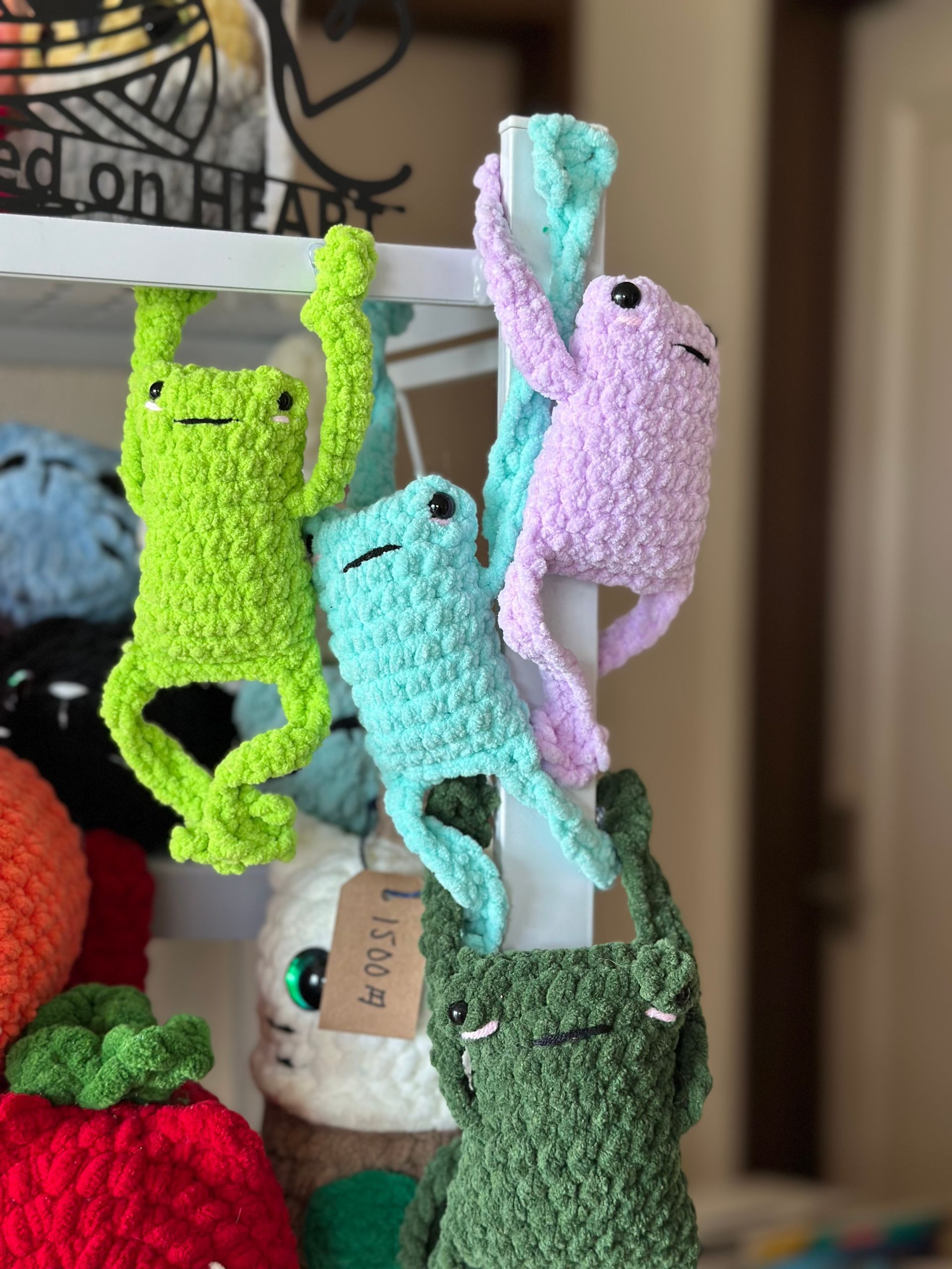 Crocheted frog toys in various colors, hanging. Yellow, pink, and green are prominent, with smiling faces and long limbs.