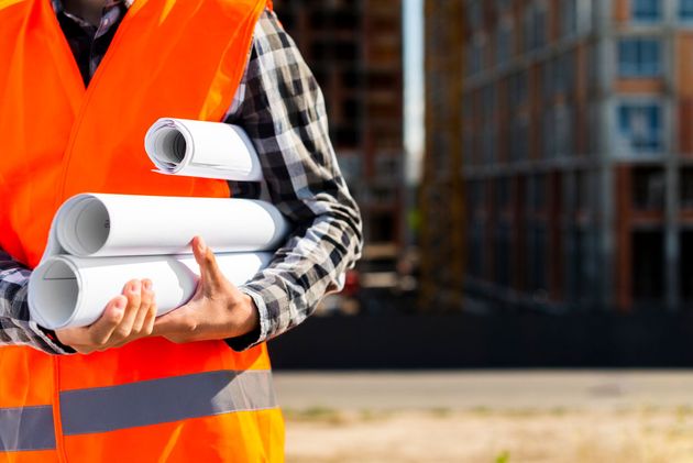 Construction worker holding rolled blueprints, wearing an orange safety vest, building in background.