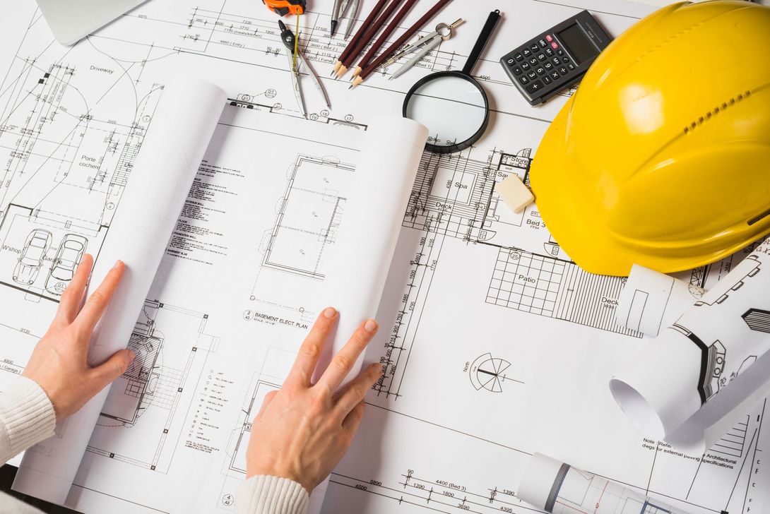 Hands examining architectural blueprints with tools, a hard hat, and calculator on a desk.