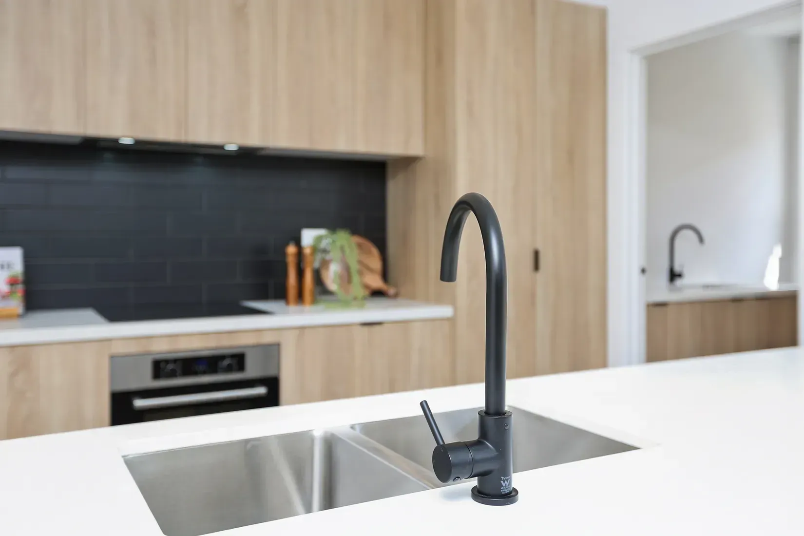 Modern kitchen with black faucet over a stainless steel sink, light wood cabinets, and black backsplash.
