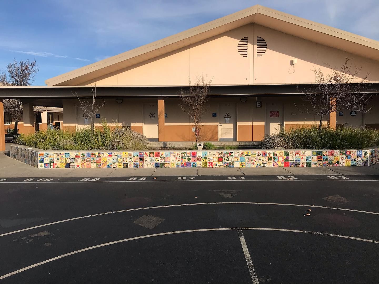 A school building exterior with a paved playground in the foreground and a long, colorful mosaic tile wall along the base.