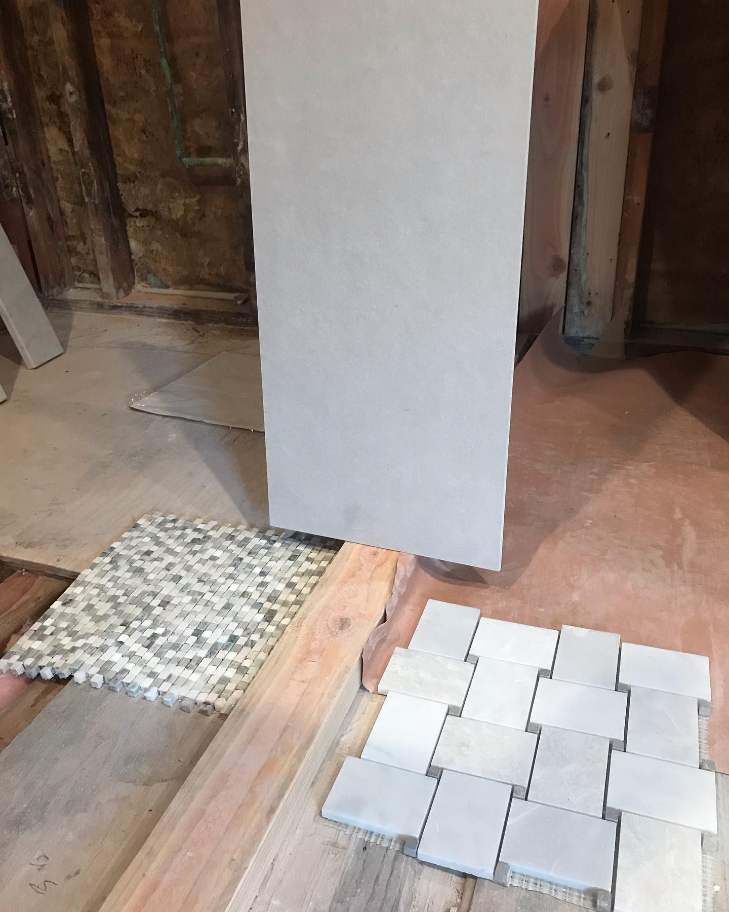 A home renovation scene showing two tile samples—one small mosaic and one basket-weave pattern—on a plywood subfloor.