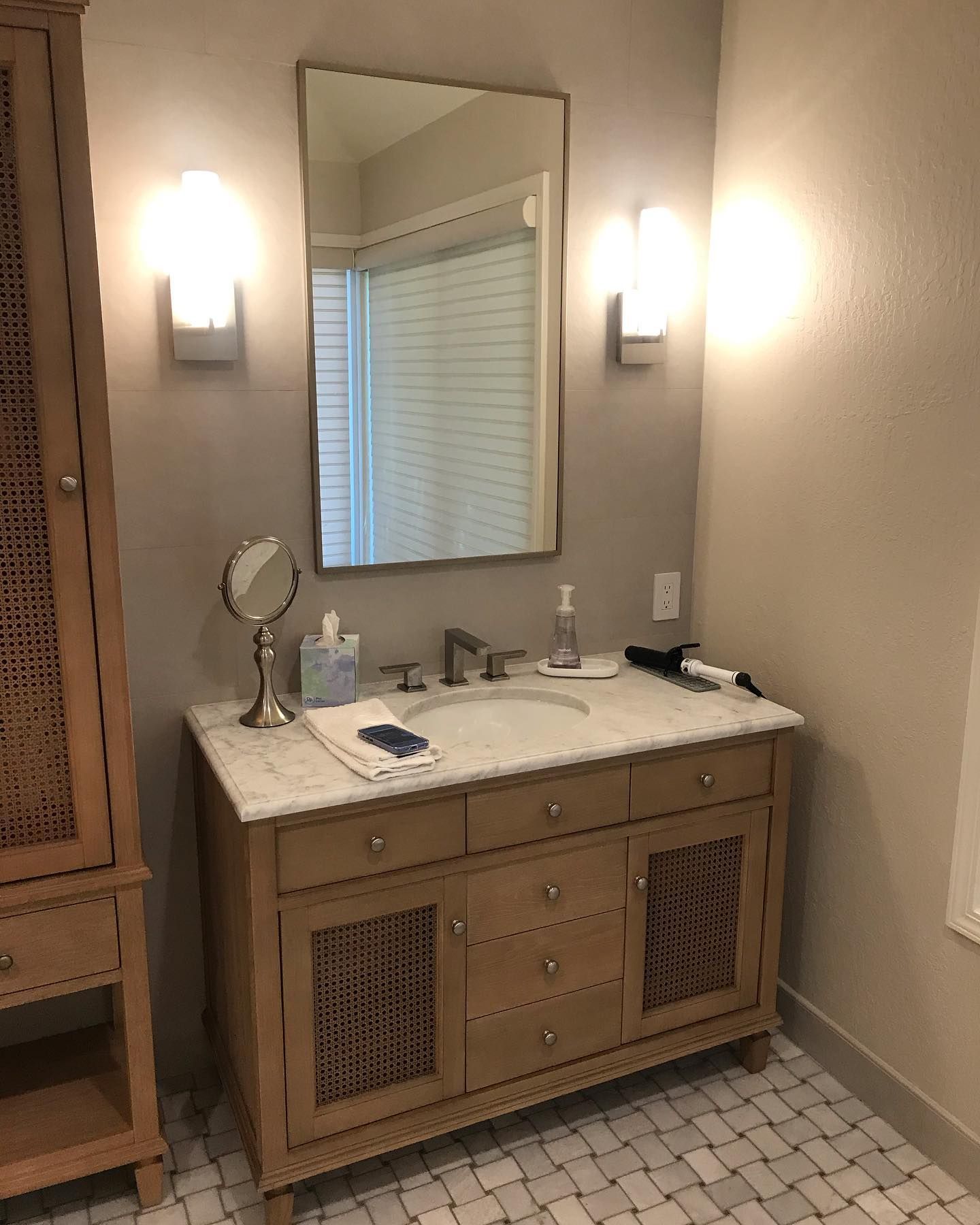 A light wood vanity with a marble countertop, mirror, and two wall sconces in a bathroom with patterned tile flooring.