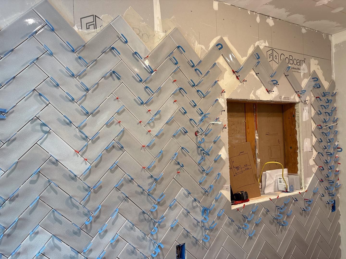 Herringbone pattern tiles installed on a wall around a window opening, featuring blue spacers between each tile.